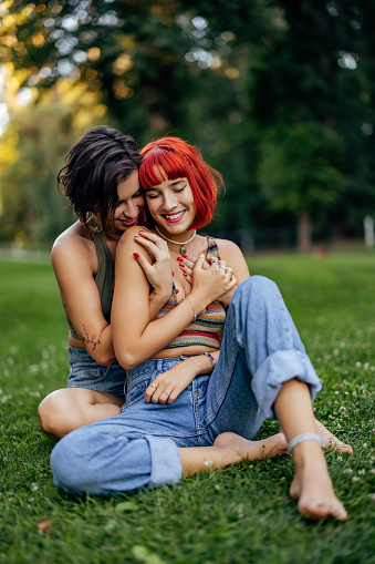 Brunette girl hugging her red hair girlfriend, sitting on the grass, enjoying nature.
