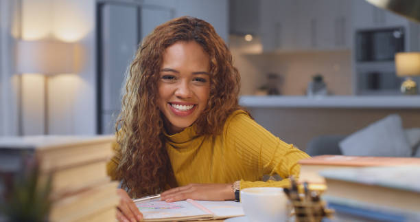 estudio, sonrisa y educación, una mujer con libros haciendo trabajo escolar en una sala de estar. inclinándose, estudiando y tomando notas, una feliz estudiante universitaria con sus tareas e investigaciones en un escritorio de su casa. - leaning forward fotografías e imágenes de stock