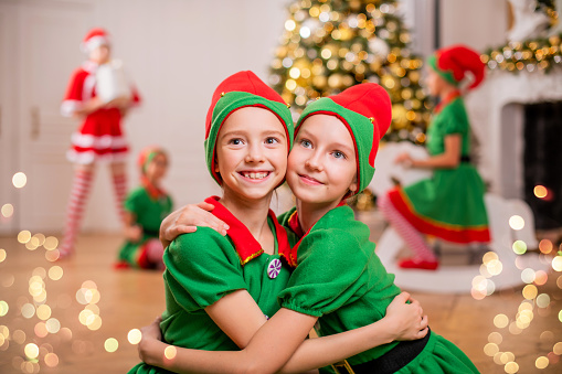 Two cheerful happy girls-friends in costumes of Santa's elves on background of Christmas tree and other children