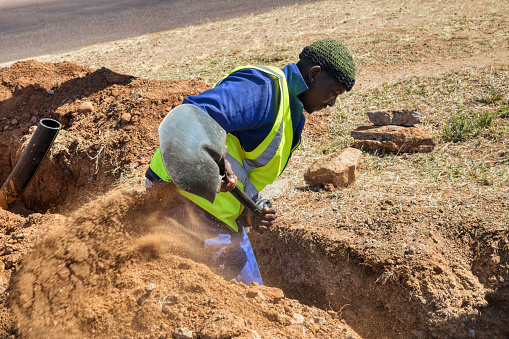 African worker in blue workwear with a beanie digging a trench in the hot sun
