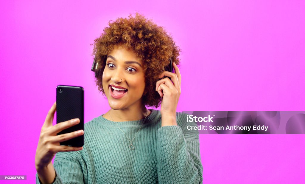 Woman with phone in hand and headphones on afro surprise smile for video call or good news via the internet. Young lady with earphones, hair curly excited by social media or music on smartphone. Headphones Stock Photo