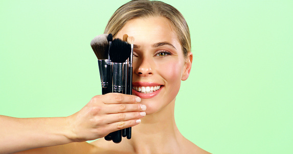 Makeup, beauty and brush with a beautiful woman in studio on a green background with mockup. Cosmetics, face and skincare with a young female holding cosmetic brushes in her hand with a smile