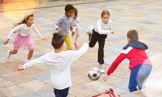 International group of sports tweenagers having fun together outdoors, playing football in school yard after lessons on autumn day