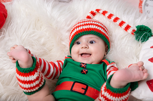 Newborn baby boy dressed in gnome costume lying on white fur carpet among christmas decorations. Christmas photo of infant in ctriped cap. New Year concept.