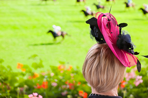 A fashionable lady, dressed for the occasion at the racetrack, watching thoroughbred race horses in a flat race on turf in front of the grandstand on a summers day. Good copy space