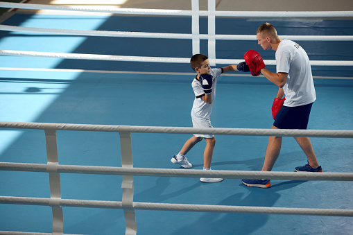 Two athletes, son and father is doing sports in boxing gym. Beginner boxer workout with his coach ar boxing ring, indoors. Sport, energy and active lifestyle. Trainer teaching kid basic techniques