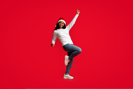 Attractive young african american woman in Santa hat and casual outfit dancing alone on red, moving her arms and legs and cheerfully smiling at camera, enjoying xmas party, copy space, studio shot