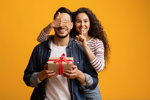 Loving Young Woman Covering Boyfriend's Eyes And Surprising Him With Gift, Cheerful Middle Eastern Lady Showing Shh Sign, Greeting Her Husband With Birthday Or Valentines Day Over Yellow Background