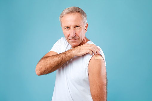 I Got My Covid-19 Vaccine. Portrait of happy senior man showing vaccinated arm after getting antiviral injection, confident male patient looking at camera posing with patch, blue studio background