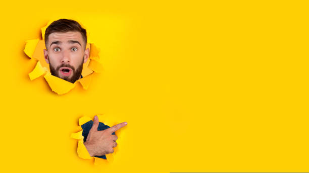 istock Surprised young caucasian guy with beard looks through hole in yellow paper and points finger to free space 1433104372