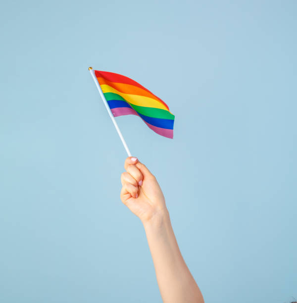 istock Closeup of a hand waving a small rainbow flag against a light blue background 1433108241