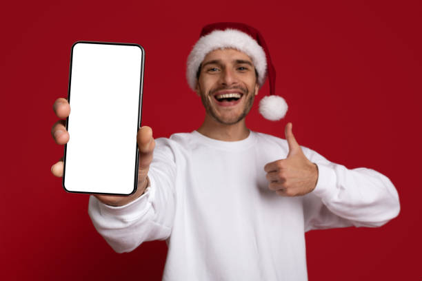 istock Cool App. Cheerful Young Guy In Santa Hat Demonstrating Blank Smartphone, Mockup 1433109829