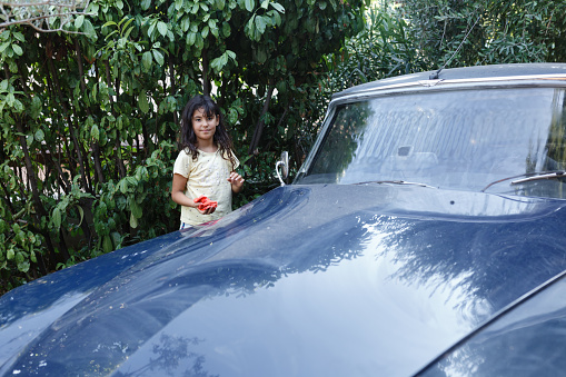 Girl with long brown hair is washing blue vintage car in the garden with orange cloth