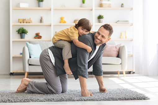 Active domestic games. Cheerful young man playing with his little son, piggybacking boy, happy kid playing cowboy on horse, smiling to camera at home interior