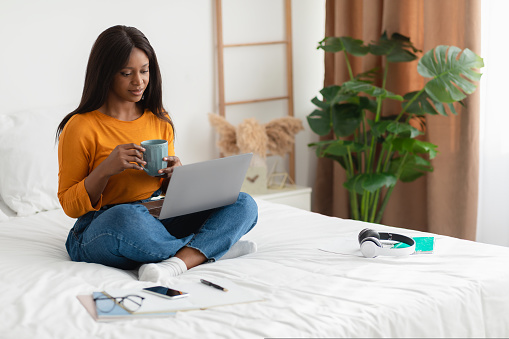 Black Woman Enjoying Coffee Using Laptop Computer Watching Film Online Sitting In Modern Bedroom At Home. Weekend Leisure, People And Technology Concept