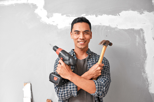 A young Asian man smiles while holding drill and hammer for home DIY improvement. High quality photo