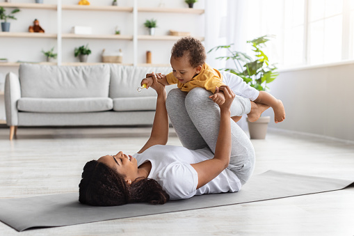 Sport Activities With Baby. Young Black Mom Exercising With Her Infant Son At Home, Happy African American Woman Balancing Baby On Knees While Training On Fitness Mat In Living Room, Copy Space