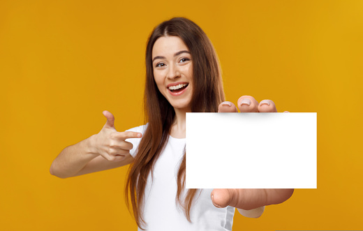 Cheerful young lady pointing at empty business card, smiling at camera on orange studio background, mockup. Cool millennial Caucasian woman presenting blank identification card
