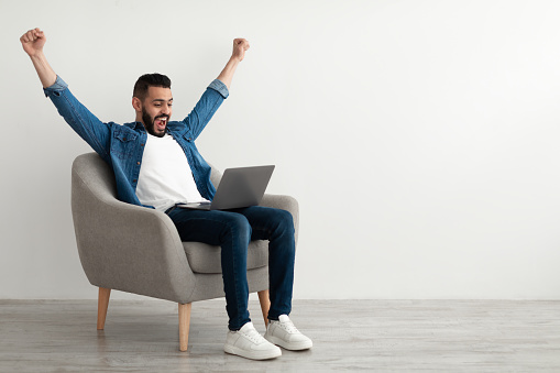 Excited young Arab man sitting in armchair with laptop, celebrating work success, winning online lottery against white studio wall, empty space. Middle Eastern guy enjoying huge sale in internet store