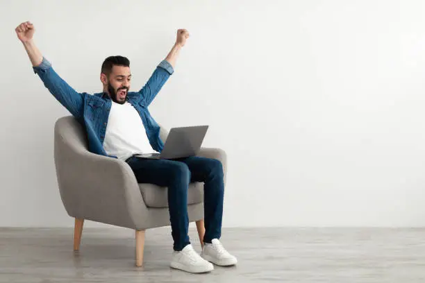 Photo of Excited young Arab man sitting in armchair with laptop, celebrating work success, winning online lottery, copy space