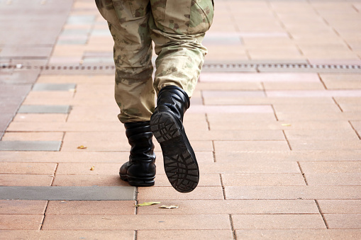 Soldado con camuflaje militar y botas caminando por la calle de la ciudad, piernas en la acera photo