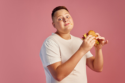 Half-length portrait of cute overweight boy in white t-shirt tasting delicious burger isolated on pink background. Fast food, taste, body positive, emotions and facial expressions. Copy space for ad