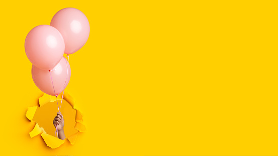 Hand of millennial african american lady holding three pink balloons through hole in yellow paper, panorama, collage, cropped. Birthday and party, celebrate and carnival, freedom, sale and advertising