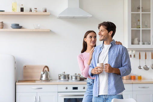 Cheerful millennial husband with cup of hot drink looks at empty space, wife hugs guy in white kitchen interior. Coffee break together, breakfast in morning and couple enjoy free time in weekend