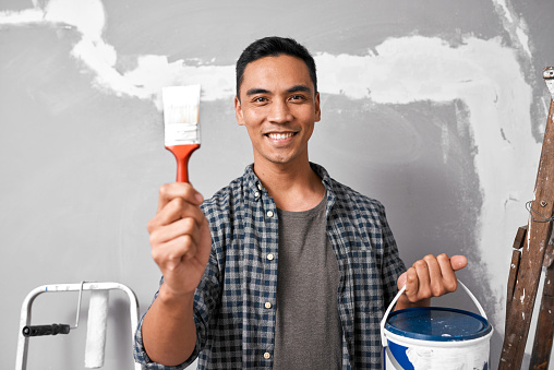 An attractive Asian man holds up paintbrush, bucket and smiles before painting. High quality photo