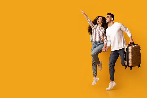 Hot Tours. Joyful Arab Couple With Suitcase Jumping And Pointing Away, Excited Young Middle Eastern Spouses Showing Copy Space On Yellow Background For Travel Offer Or Advertisement, Full Length