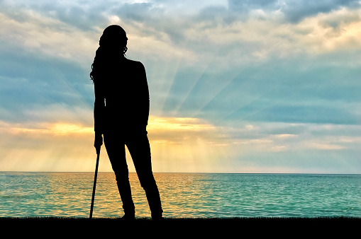 A silhouette of a woman supports herself with a crutch admiring the sea sunset. Concept of people in rehabilitation