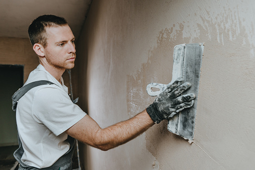 Male builder in work overalls plastering a wall using a construction trowel. Horizontal panorama banner with blank space for text.
