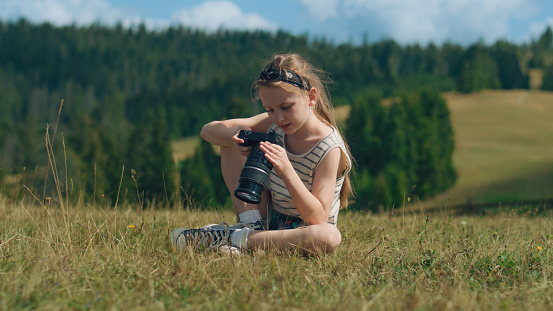 Young photographer watching results of work, sitting on mild grass hill. Girl relaxing and feeling warm wind currents, spending leisure time outdoor