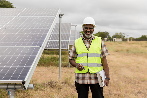 The mature adult male engineer, standing by the solar panels installed in the field, smiles for the camera.