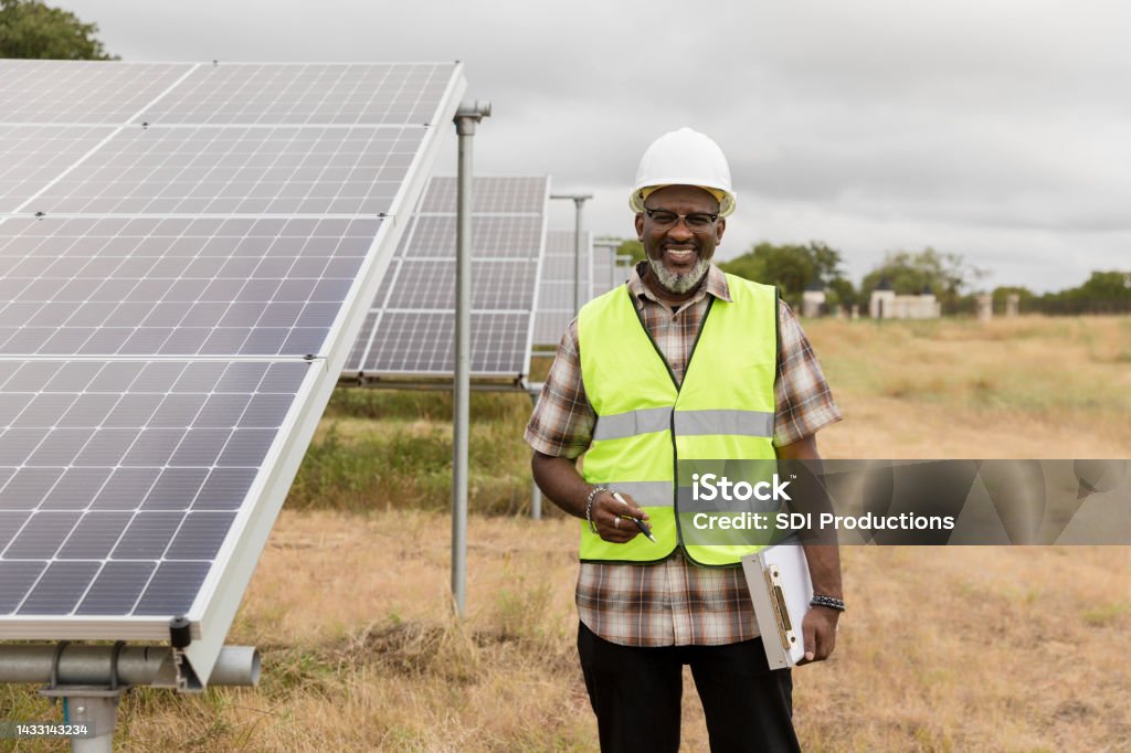 Male engineer stands by solar panels in field The mature adult male engineer, standing by the solar panels installed in the field, smiles for the camera. Maintenance Engineer Stock Photo