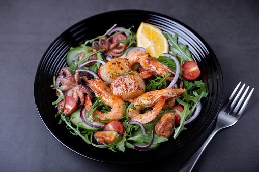 Salad with seafood (shrimp, scallops, octopus, mussels), arugula, tomatoes, cucumbers, red onions and lemon on a black plate. Black background, close-up, selective focus.