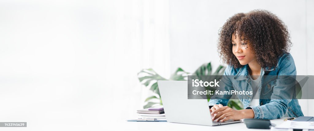 American teenage woman sitting in white office with laptop, she is a student studying online with laptop at home, university student studying online, online web education concept. Using Computer Stock Photo
