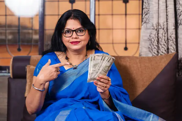 Young Indian woman holding currency notes while sitting on sofa at home.