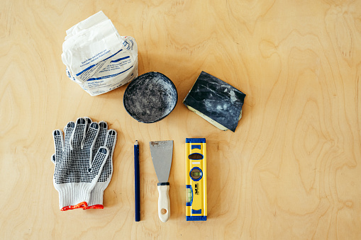 Top view of wall repairing tools on hardwood floor. Flat lay of putty knife, hand gloves, wall putty packet, bowl, sand paper, pencil and spirit on wooden floor.