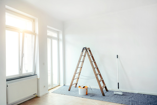 Empty apartment after renovation. Painting in a white room with windows with a stepladder, paint roller and paints on the floor.