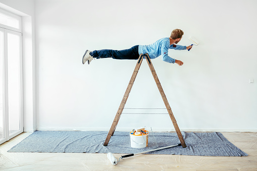 Young man balancing on a ladder and painting living room wall. Man being silly while renovating his home.
