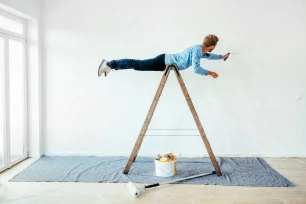 Photo of Young man balancing on a ladder and painting living room wall