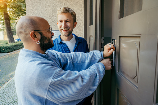 Two young male friends opening the front door of their apartment building and smiling. Men unlocking the new house door with key and smiling.