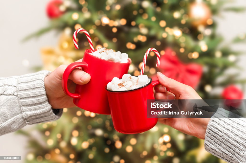 two red mugs with a hot drink and marshmallows the hands of a man and a woman hold two red mugs with a hot drink and marshmallows on the background of the Christmas tree Christmas Stock Photo