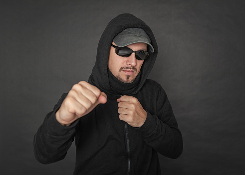 Man in the black hoody with hood wearing sunglasses with fist up ready to fight on dark background