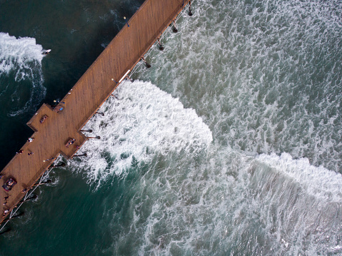 A Dramatic Shot of a Pier Off of the Coast of California, A dark and gloomy day near a pier in southern California, Straight Down Drone Shot of a Pier with Waves Crashing around it.