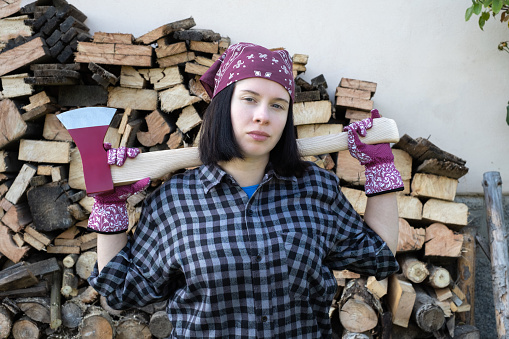 Young adult woman wearing a bandana, stands and looks at the camera, holding a chopping ax behind her shoulders against the background of prepared firewood. Strong woman ready for an energetic crisis.