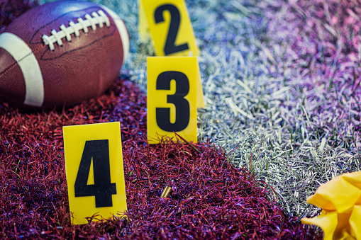Close-up pf a crime scene marker in the end Zone of an American football game, with an American football and evidence markers. Along with a referee’s hat and yellow penalty flags.