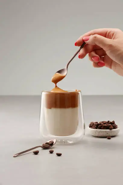 Cup with Dalgona coffee. Iced fluffy creamy whipped trend drink with coffee foam and milk in a glass with double glass. Trendy Korean whipped coffee. A woman's hand takes foam from a cup of Dalgona coffee with a spoon. Fluffy drink. Shallow depth of field