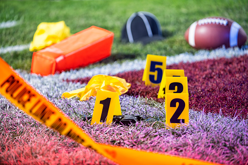 A crime scene in the end Zone of an American football game, with crime scene tape and evidence markers. Along with a referee’s hat and yellow penalty flags.